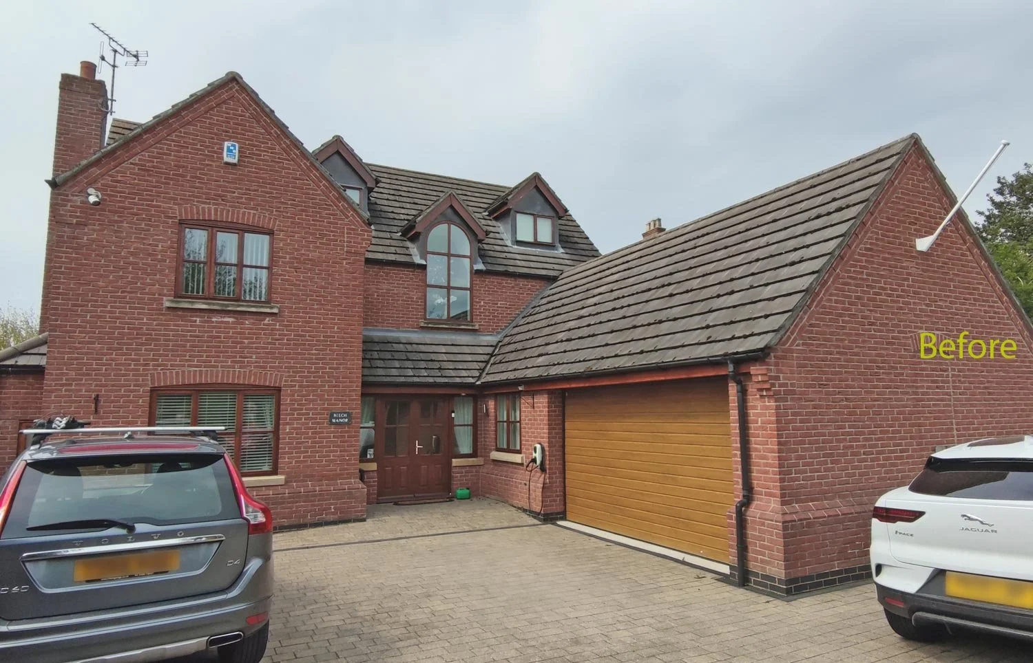 A brick house with multiple gabled roofs, a garage with a wooden door, and two cars parked in the driveway. The house has several windows and a front door, with a small sign that says 'BEUCH MANOR'. The word 'Before' is written in yellow text on the 