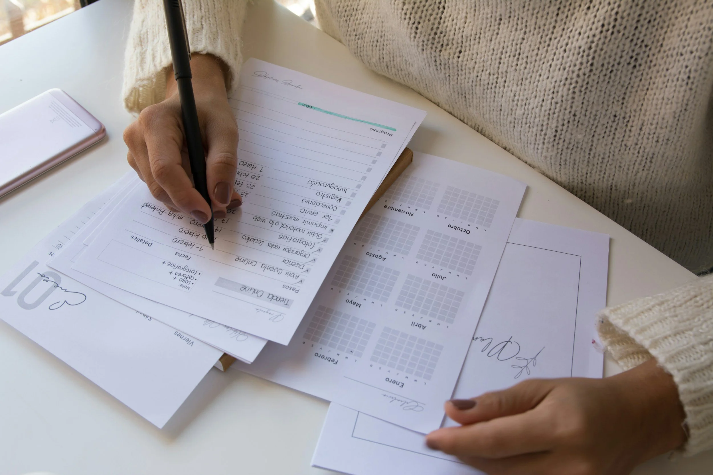 A person wearing a cream knit sweater is filling out a planner with a black pen. The planner is on a white table with scattered sheets of paper and a pink smartphone nearby.
