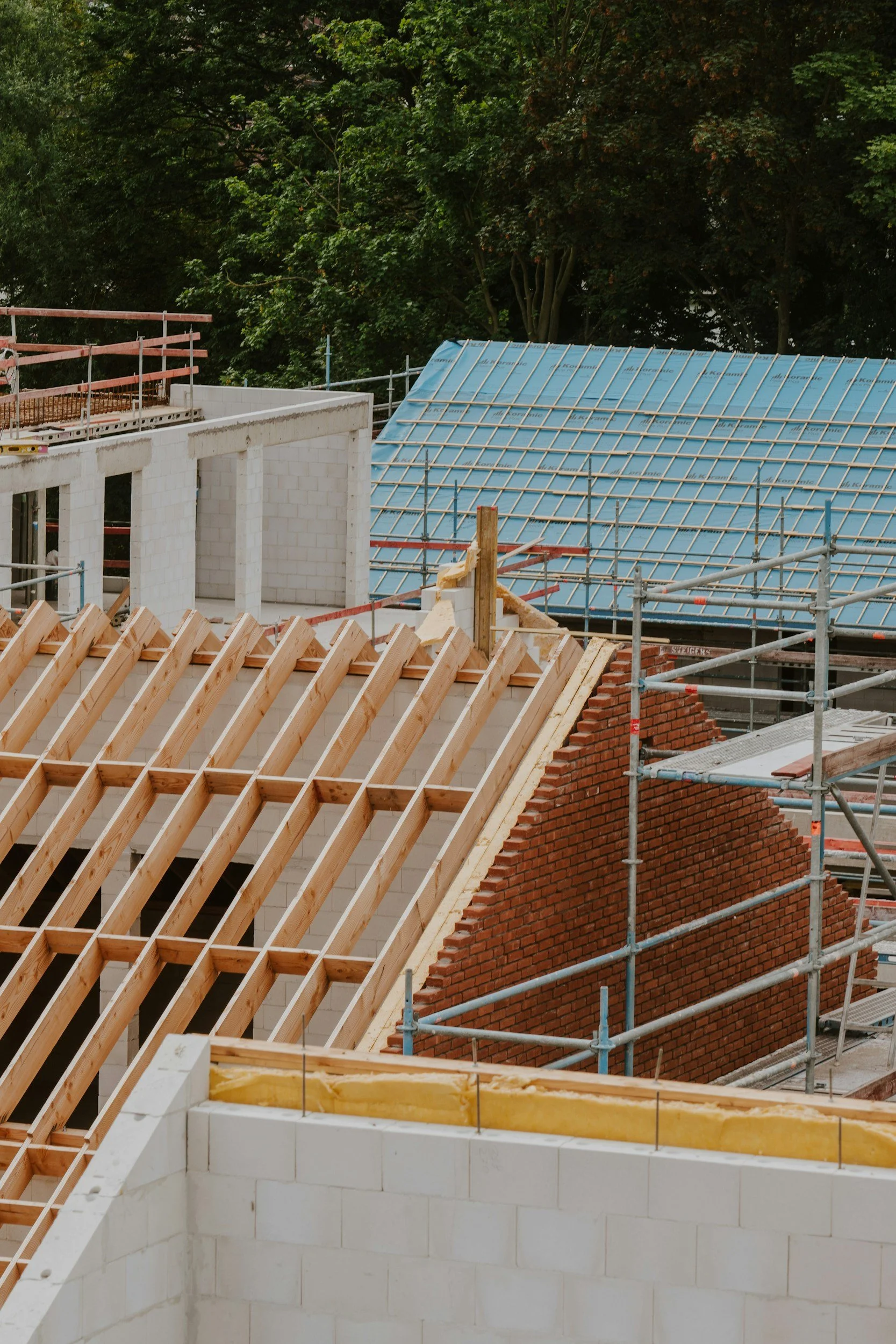 Construction site with partially built white block walls, wooden roof framing, and a brick wall, set against a background of dense green trees.