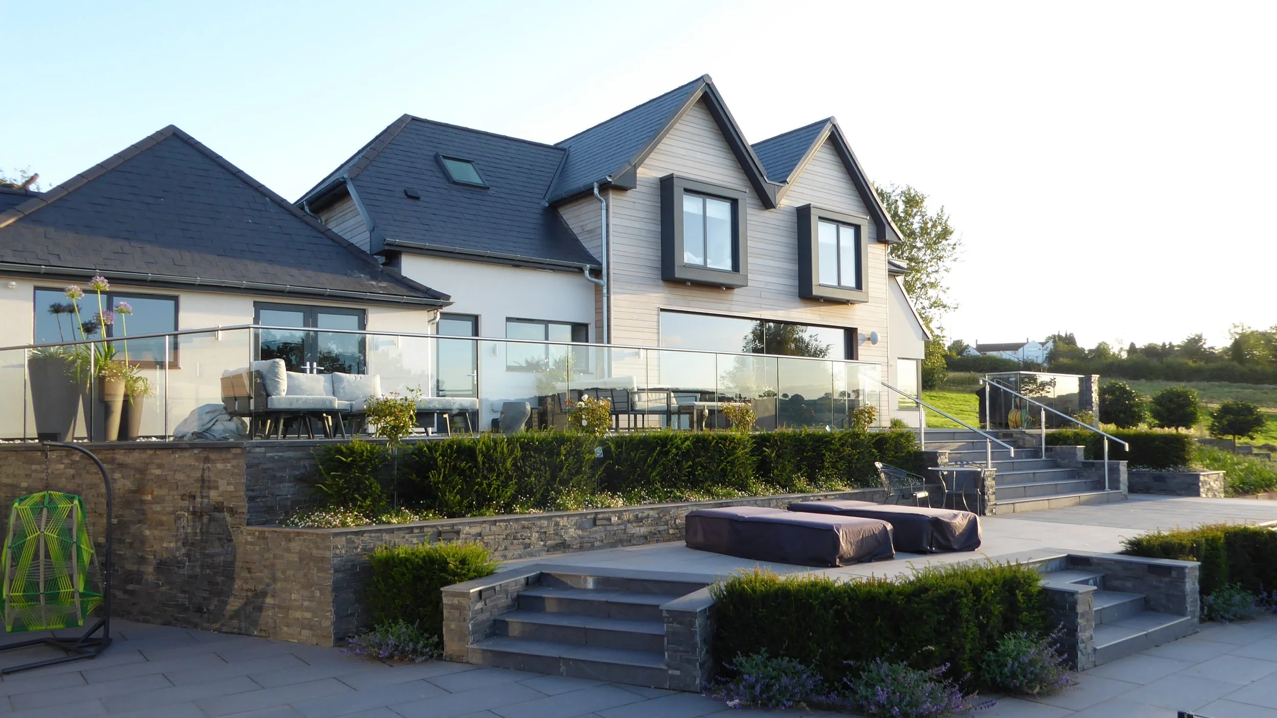 Modern two-story house with a dark roof, large windows, and a balcony with outdoor furniture; landscaped backyard with steps, hedges, and potted plants, under clear sky.