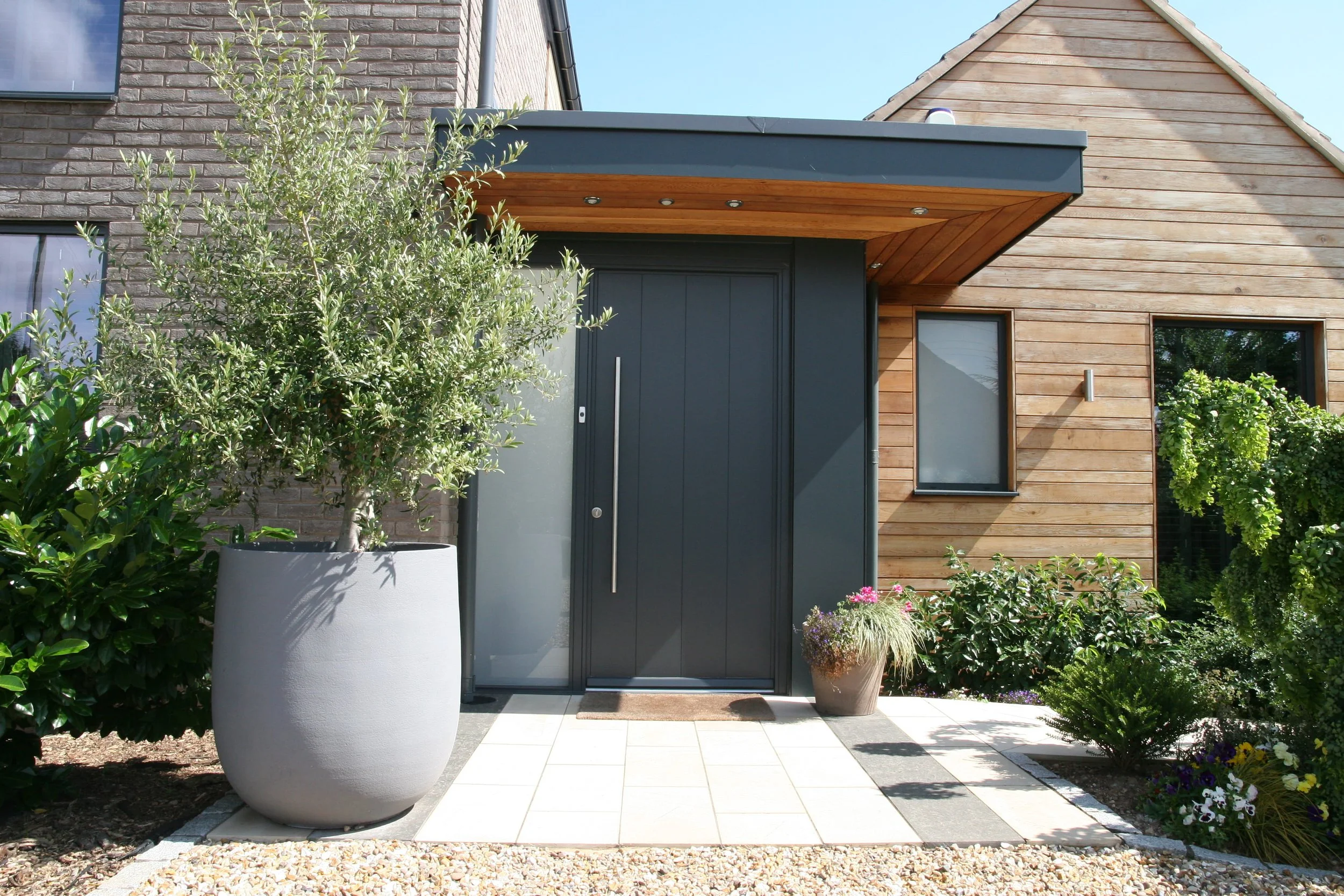 Modern house entrance with dark gray door, wooden overhang, and surrounding greenery including a large potted tree and flowering plants.