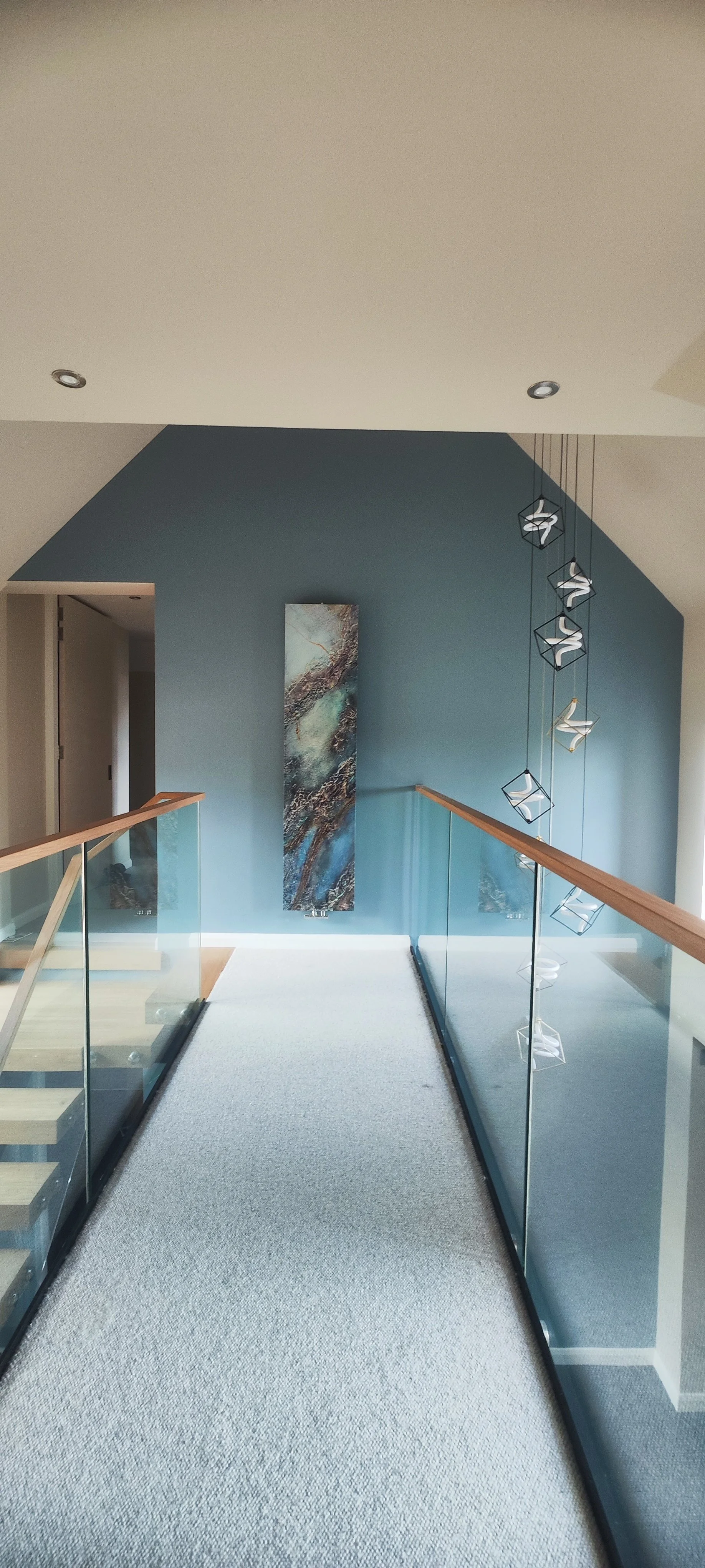 Interior hallway with a blue accent wall, abstract artwork, glass railing, wooden handrails, and modern hanging light fixtures.