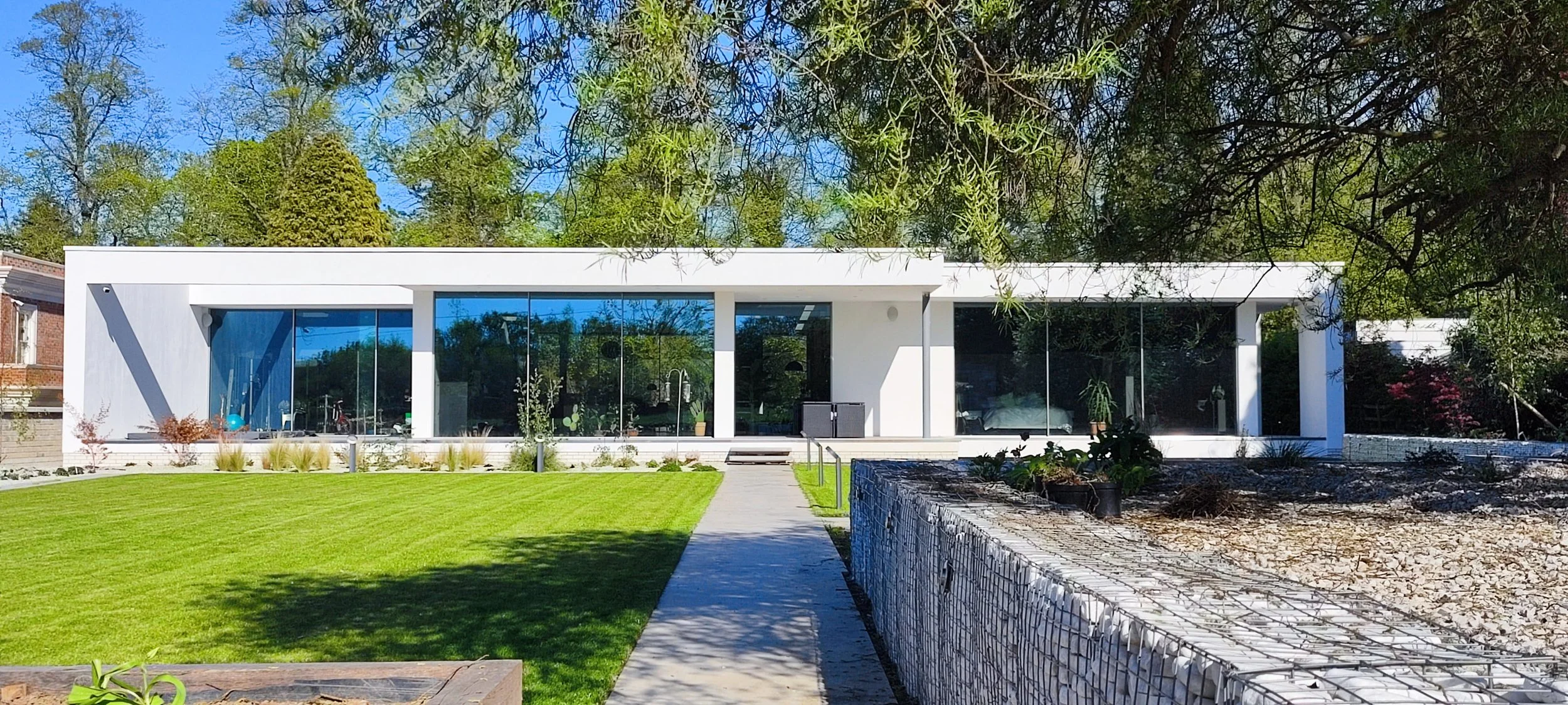 Modern white house with large glass windows, a green lawn, and a concrete pathway leading to the entrance, surrounded by trees and landscaping under a clear blue sky.