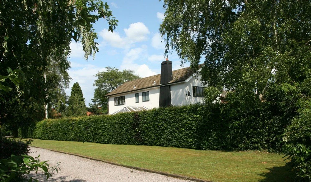 A white two-story house with a dark chimney, surrounded by trees and a tall hedge, under a partly cloudy sky.