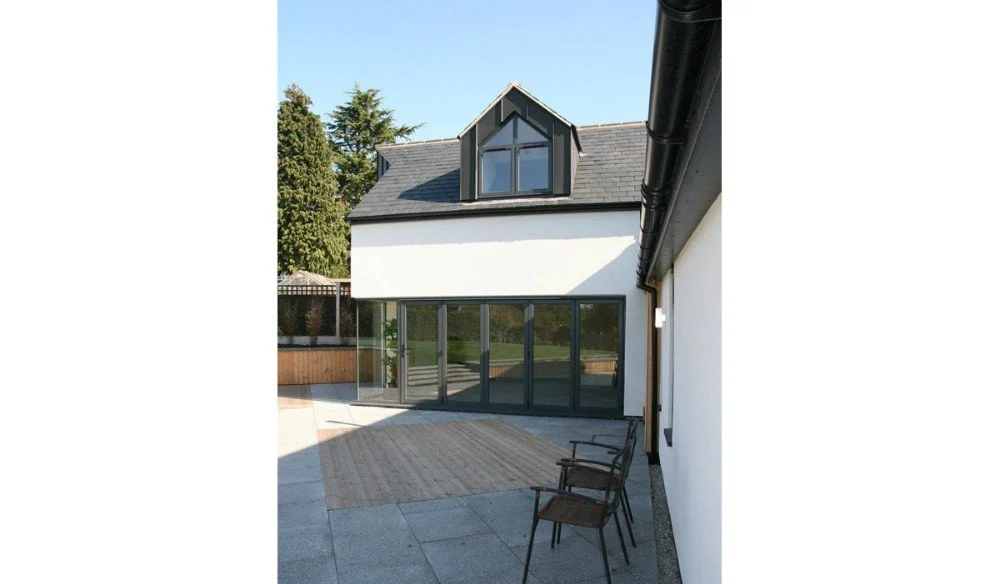 Modern house exterior with a large glass sliding door, white walls, a small front porch, and two chairs on the patio.