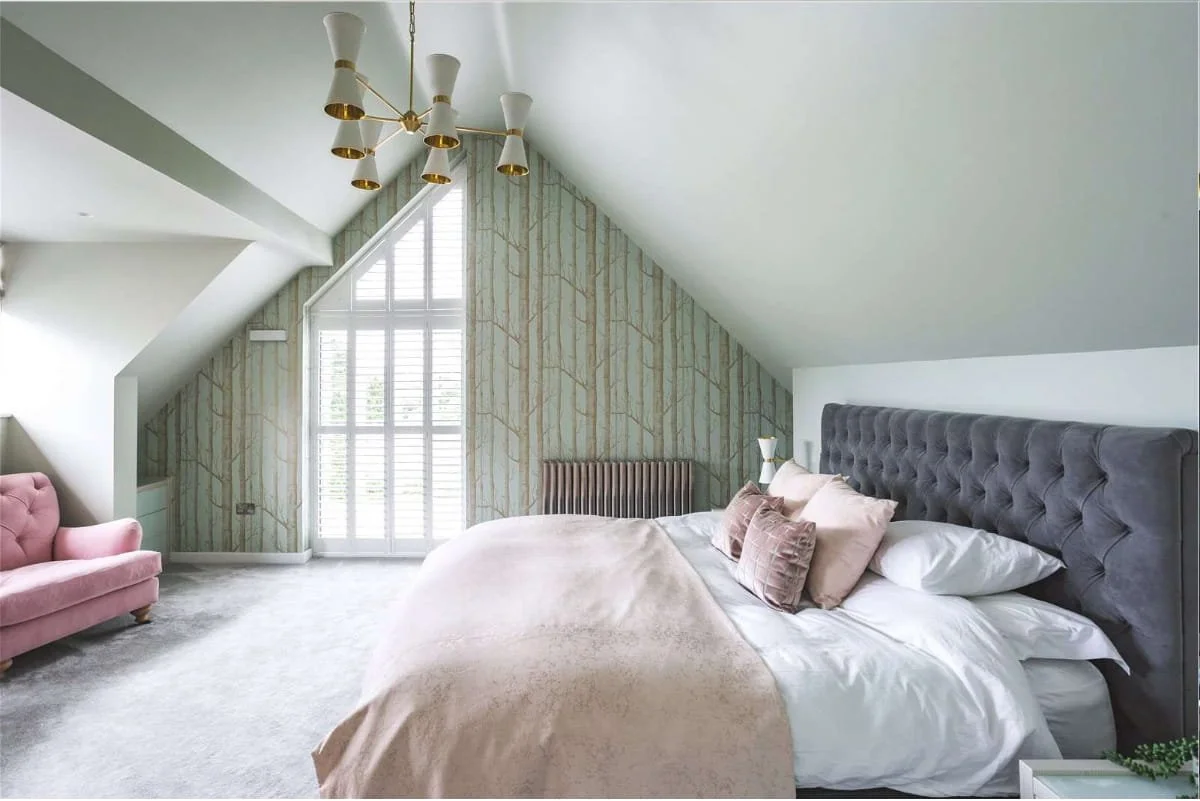 A bedroom with a sloped ceiling, large bed with gray headboard, pink and beige pillows, white bedding, a pink armchair to the left, a decorative wallpaper behind the bed, a window with shutters, and a modern chandelier hanging from the ceiling.