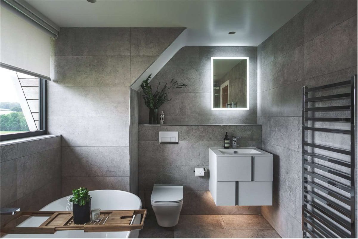 Modern bathroom with gray tiled walls, a white bathtub with a wooden tray and potted plant, a wall-mounted toilet, a white vanity with a sink, a backlit mirror, a towel warmer, and a window with a view of trees outside.