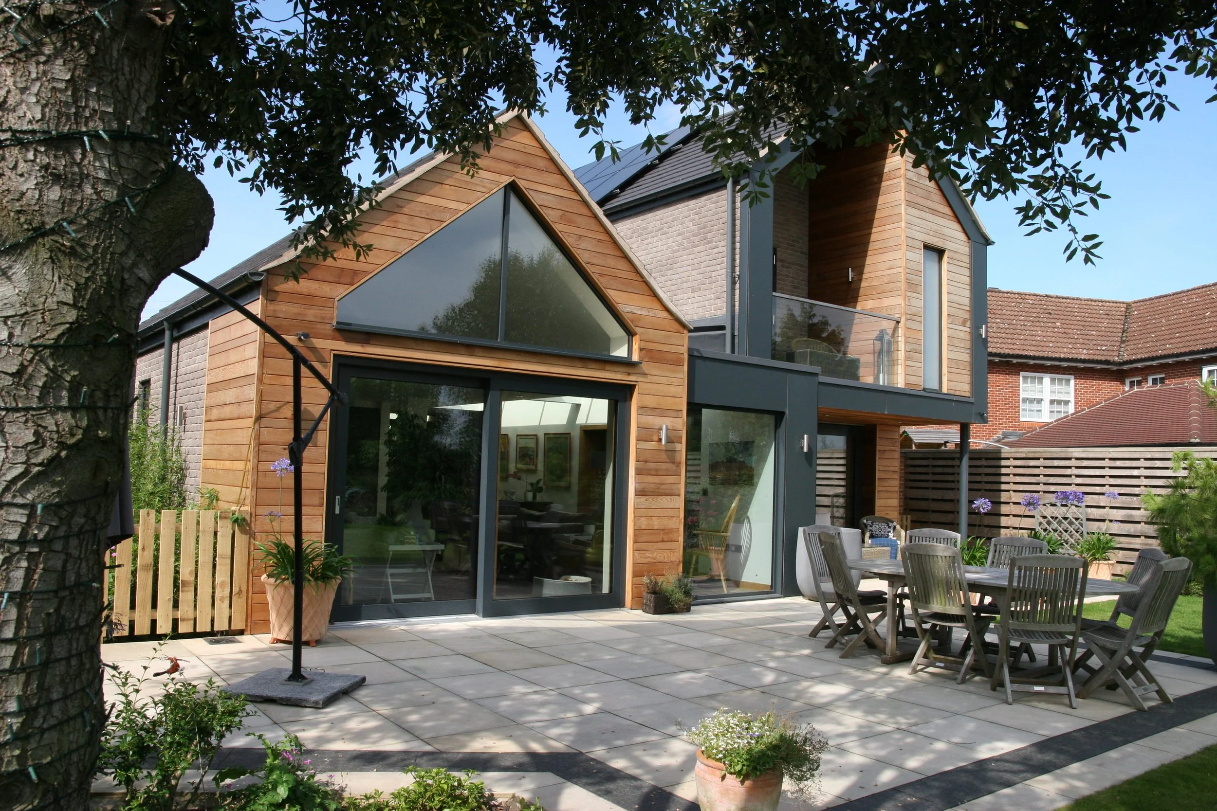 Modern house with wooden and brick exterior, large glass windows, and a patio with outdoor furniture and potted plants, surrounded by a garden with trees and flowers.