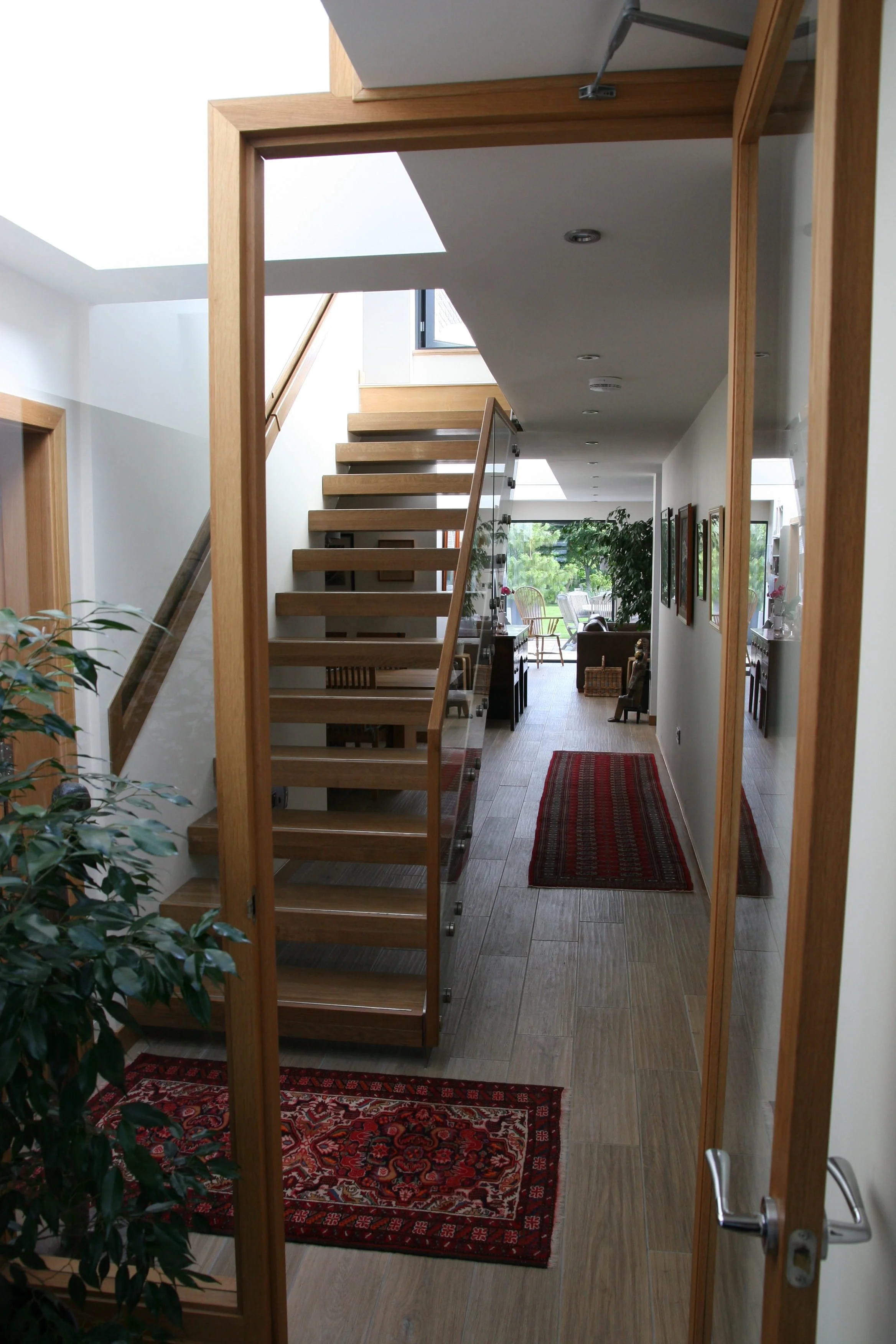 View through a glass door into a modern, bright living space with a staircase, hardwood flooring, a red patterned rug, framed pictures on the wall, and large windows with greenery outside.