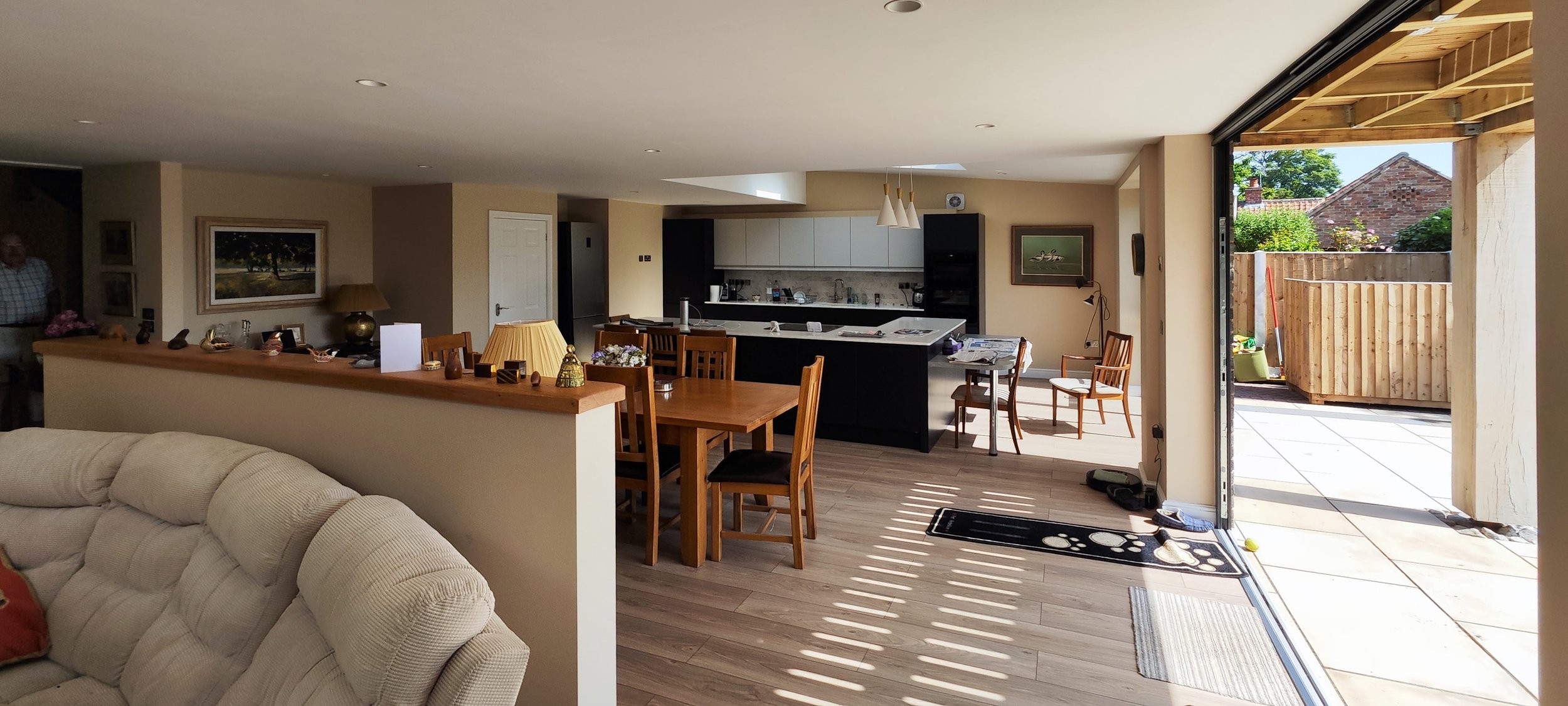 Open-concept living space with a kitchen, dining area, and outdoor patio, featuring wooden furniture, a beige sofa, and sunlight streaming in.