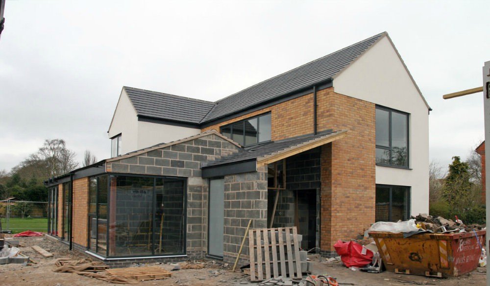 A two-story house under construction, made of brick and white stucco, with large windows and a black roof.