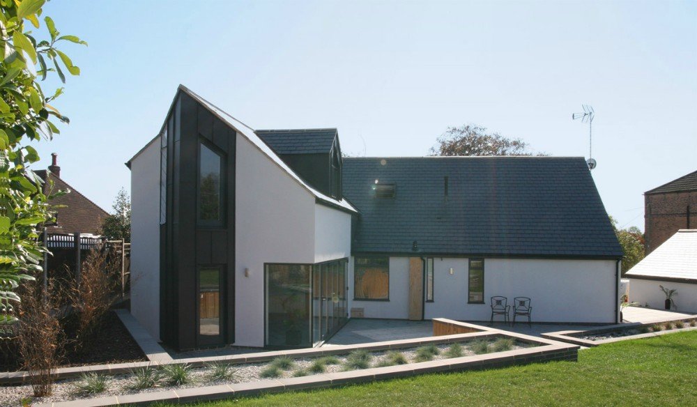 Modern house with black and white exterior, large windows, and a small outdoor seating area, surrounded by greenery and neighboring houses.