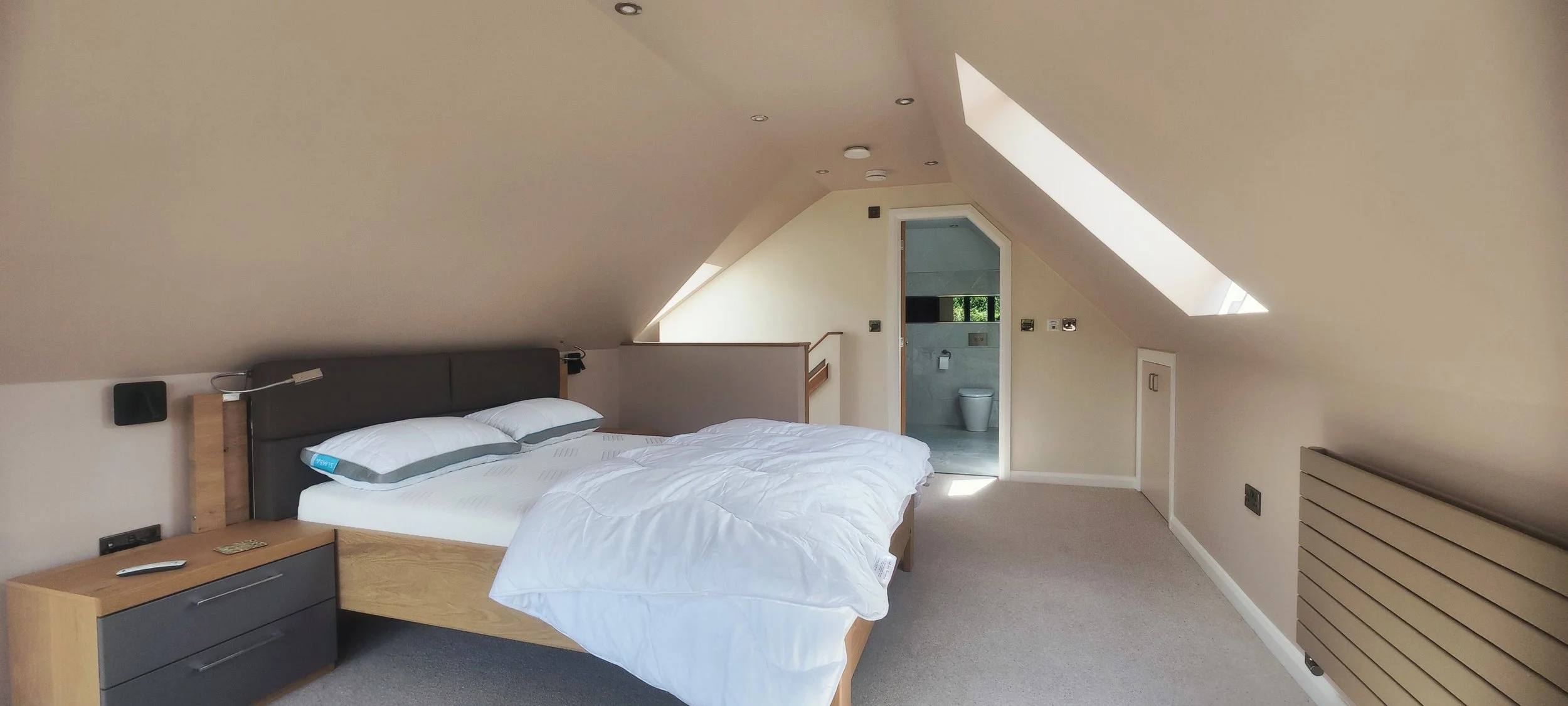 A cozy attic bedroom with a sloped ceiling, a large bed with white bedding, a bedside table with a lamp, and a view into a small bathroom through an open door.