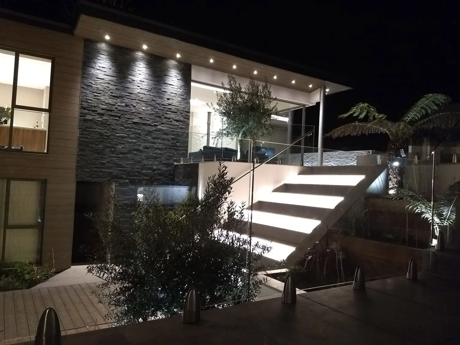Nighttime view of a modern house exterior with illuminated outdoor staircase, glass railing, stone accent wall, and lush landscaping.