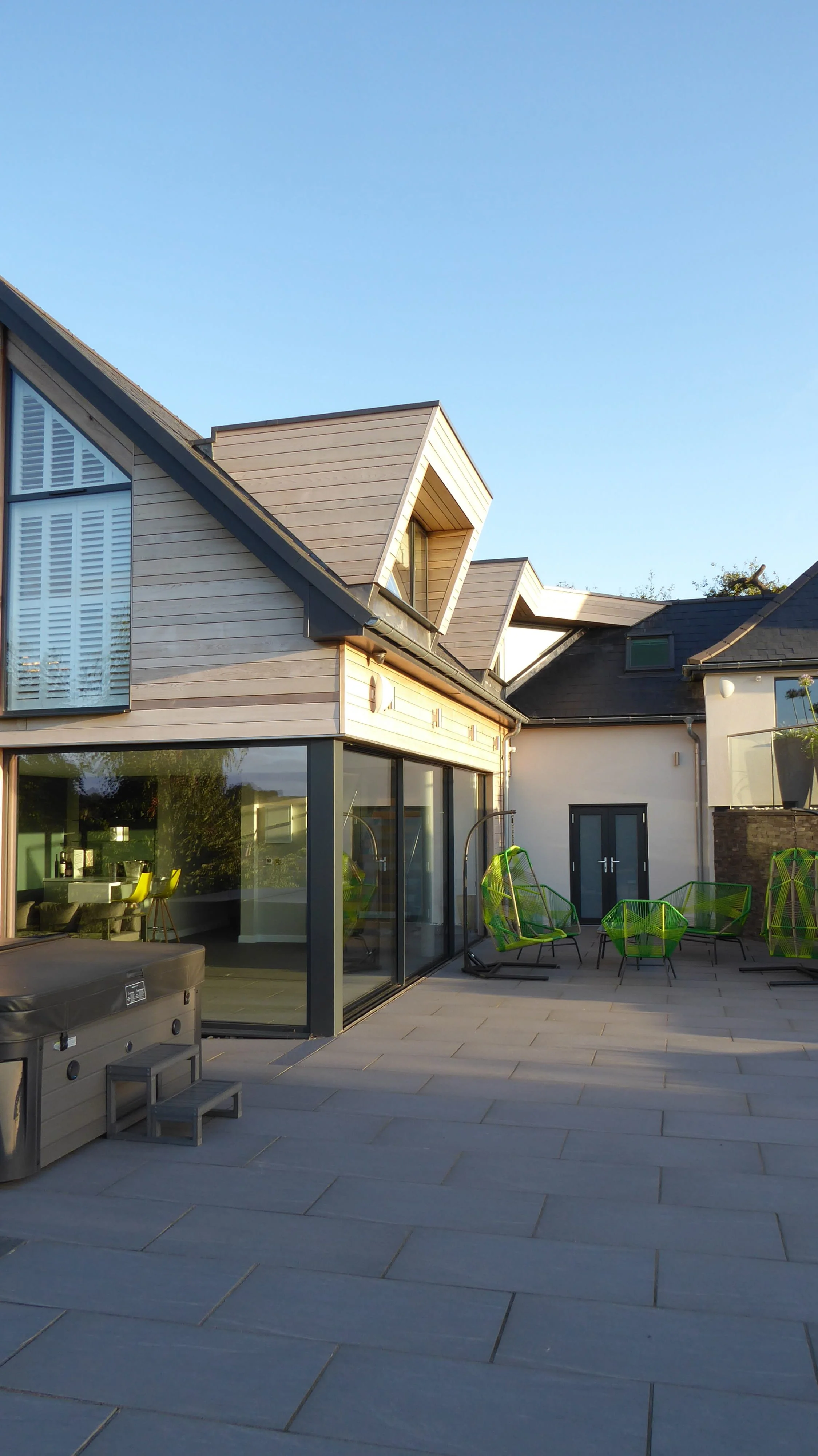 Modern house with a patio featuring green outdoor chairs, large windows, and a wooden and glass exterior, under a clear blue sky.