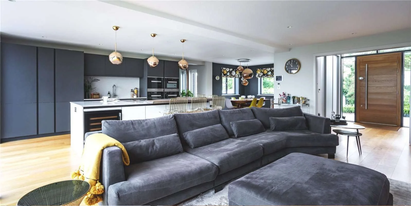 Open-concept living room and kitchen with gray and white color scheme, pendant lights, black cabinetry, a sectional sofa, and wooden flooring.