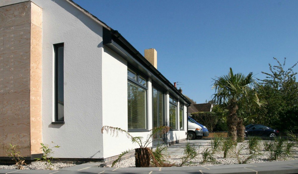 Modern house exterior with white textured walls, vertical window, large glass windows, parked cars, palm tree, and landscaped yard.