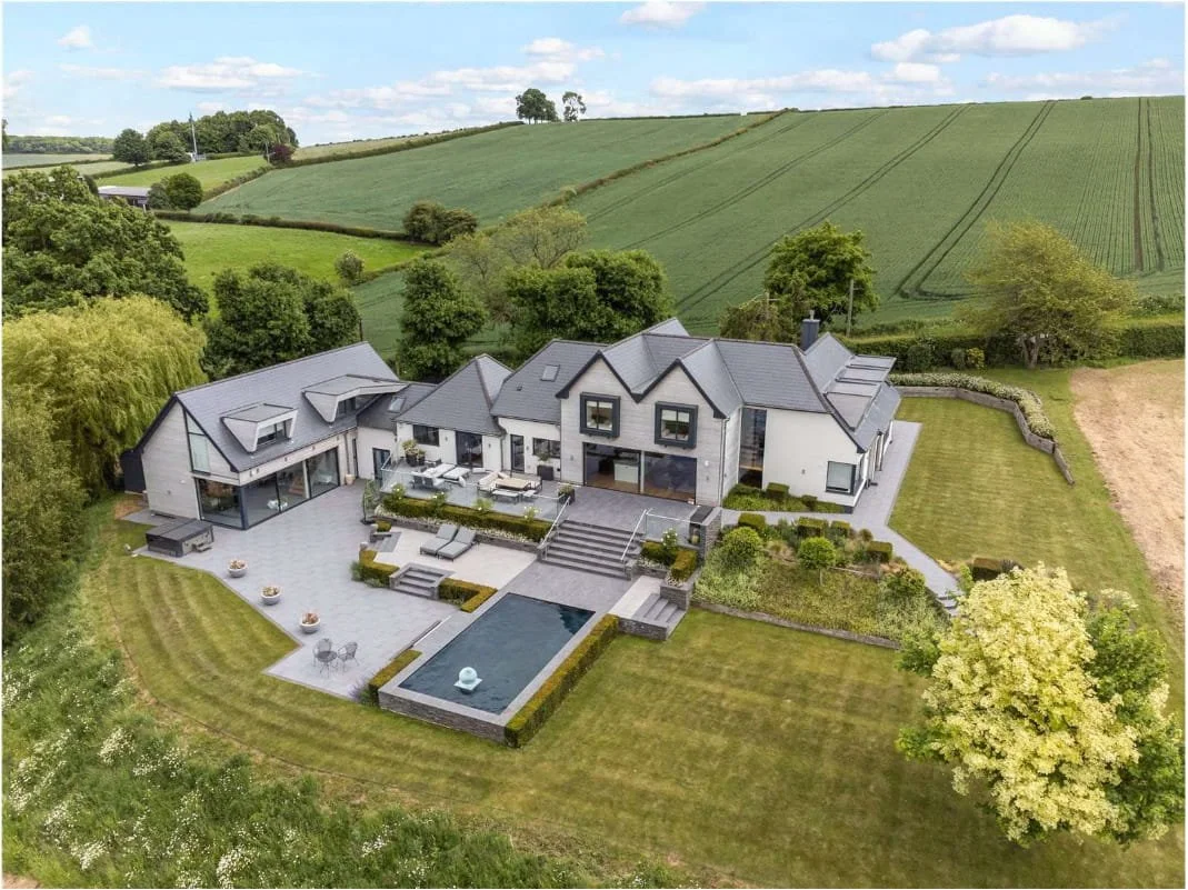 A modern luxury house with multiple sections, a swimming pool, and a landscaped yard surrounded by green fields and trees.