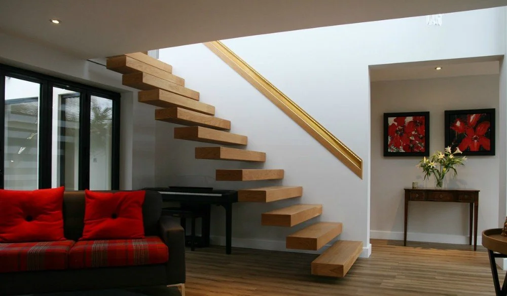 Interior view of a modern living room with a floating wooden staircase, a black sofa with red cushions, a small wooden table with flowers, and framed artwork on the wall.