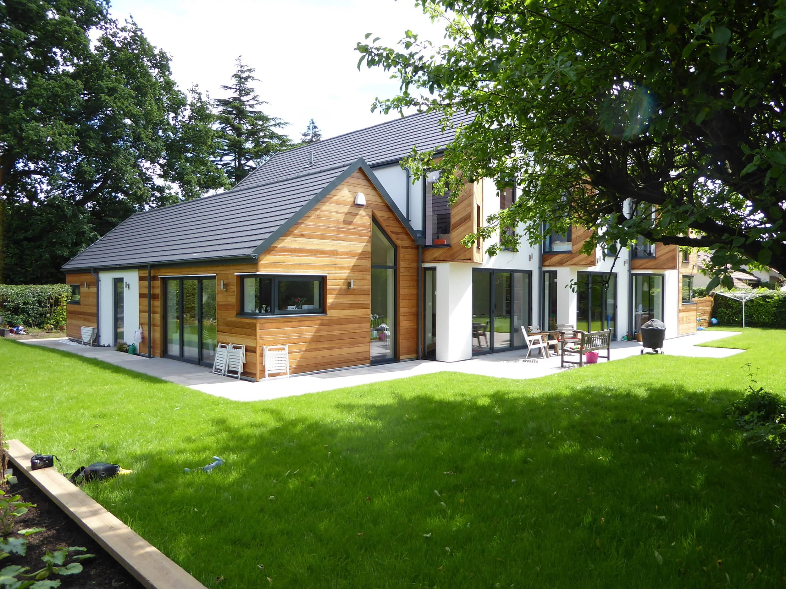 Modern house with wooden and white exterior, large glass windows and doors, surrounding green lawn, trees, and outdoor furniture, under a partly cloudy sky.