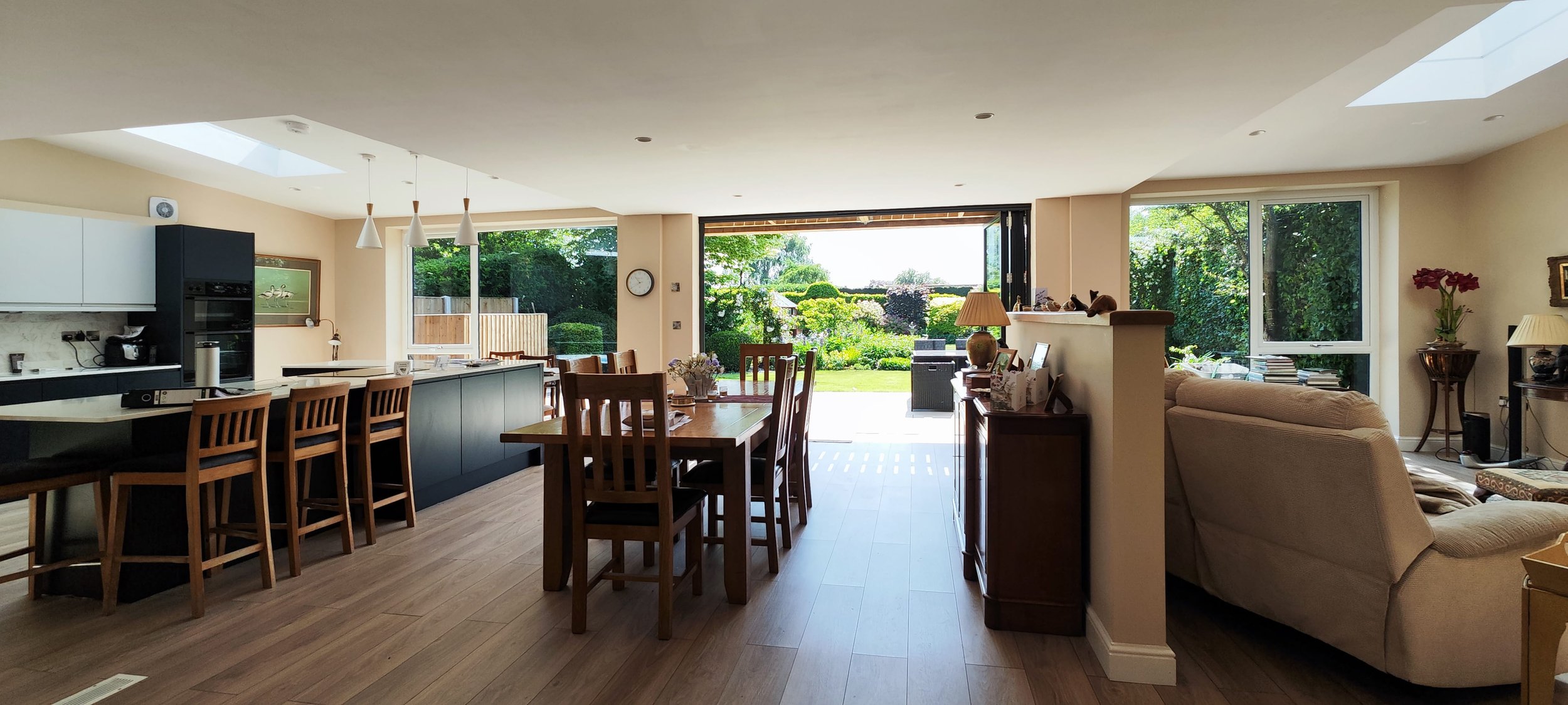 A spacious open-concept living and dining area with large windows and sliding glass doors opening to a green backyard. The room includes a kitchen island with barstools, a dining table with chairs, and a cozy sitting area with a sofa and armchair. Na