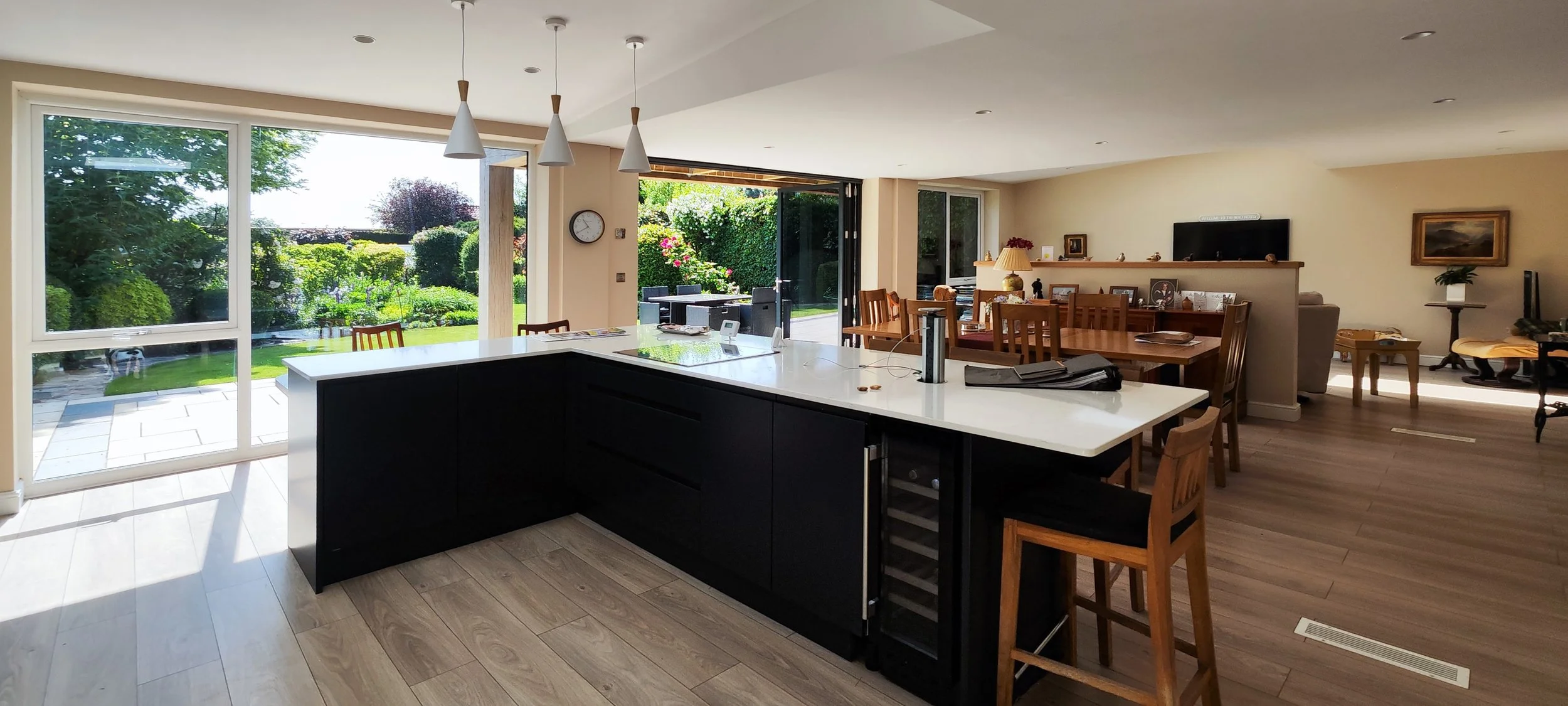 Open-concept living space with large windows showing a lush green backyard, oak cabinets and a white countertop, dining area with multiple wooden chairs, and a sitting area with a television, bookshelf, and artwork.