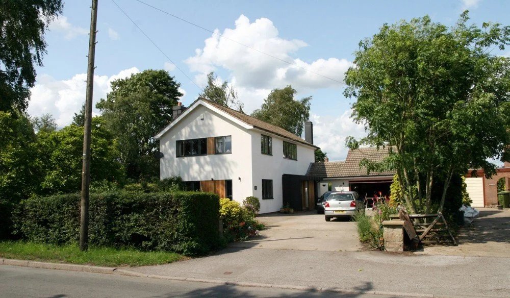 A white two-story house with a brown roof, surrounded by lush green trees and shrubs, with a driveway leading to a garage and a silver car parked outside, under a partly cloudy sky.