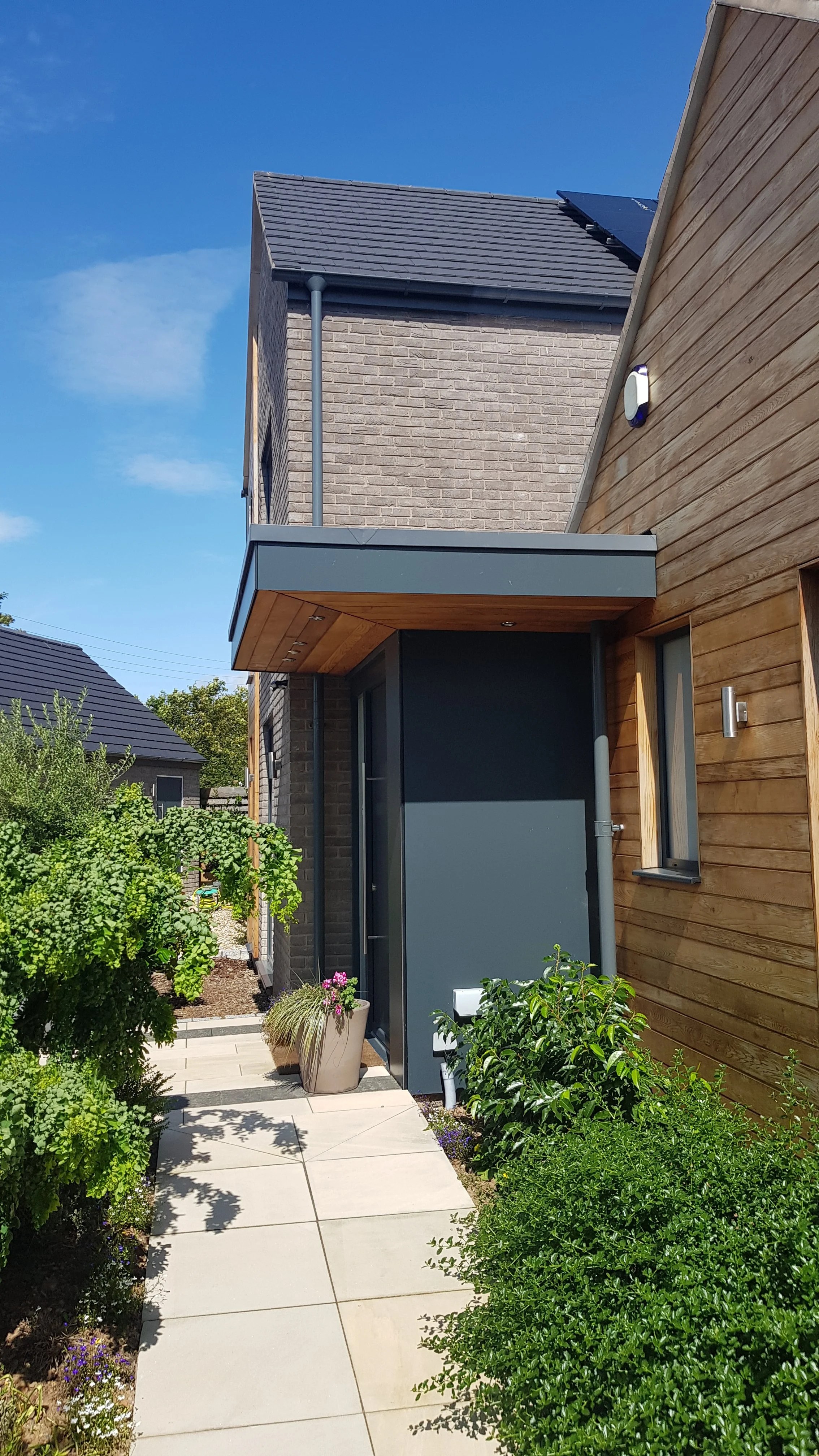 Modern house with a brick and wooden exterior, small front garden with potted plants, and a paved walkway.