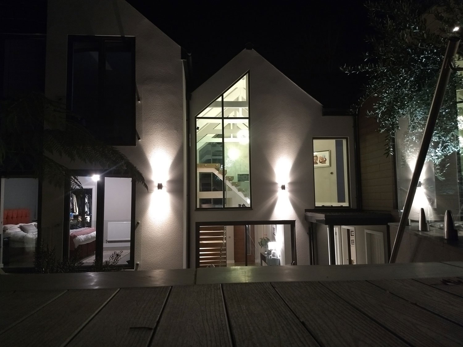 Nighttime view of a modern home with illuminated exterior lights, large windows showing interior stairs and rooms, and wooden deck in the foreground.