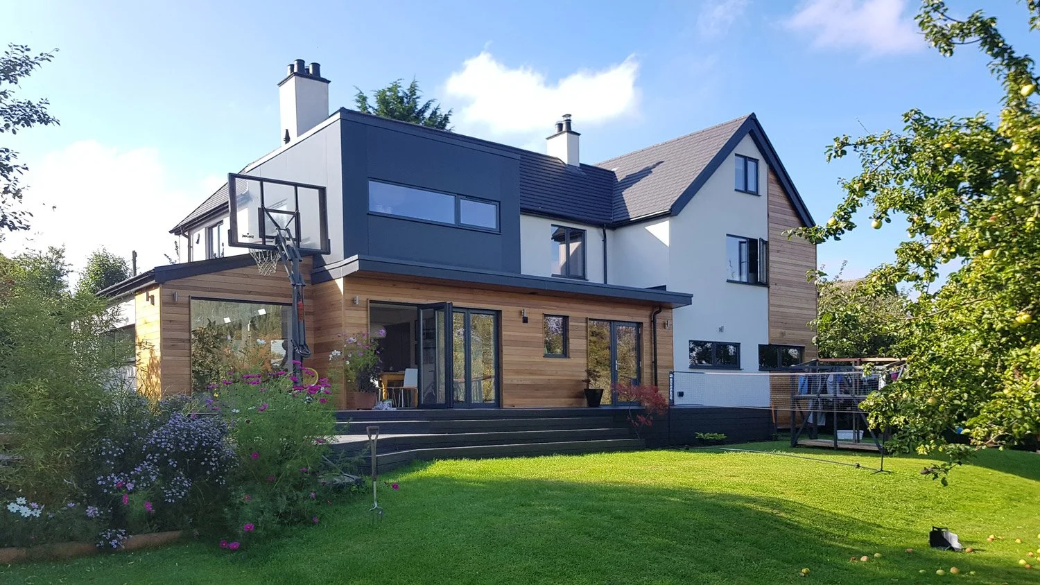 Modern multi-story house with a wooden and white exterior, large windows, and a backyard with a grassy lawn and garden.