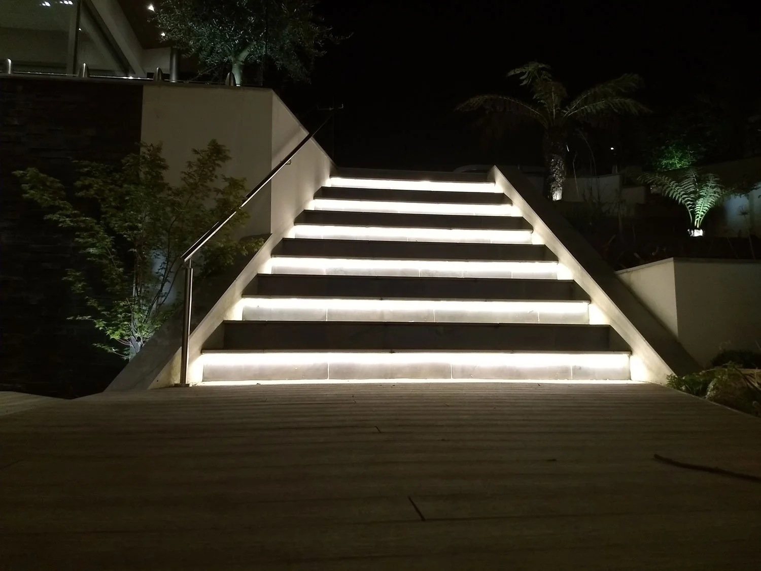 Nighttime outdoor staircase with illuminated steps, metal railing, surrounding plants, and trees in the background.