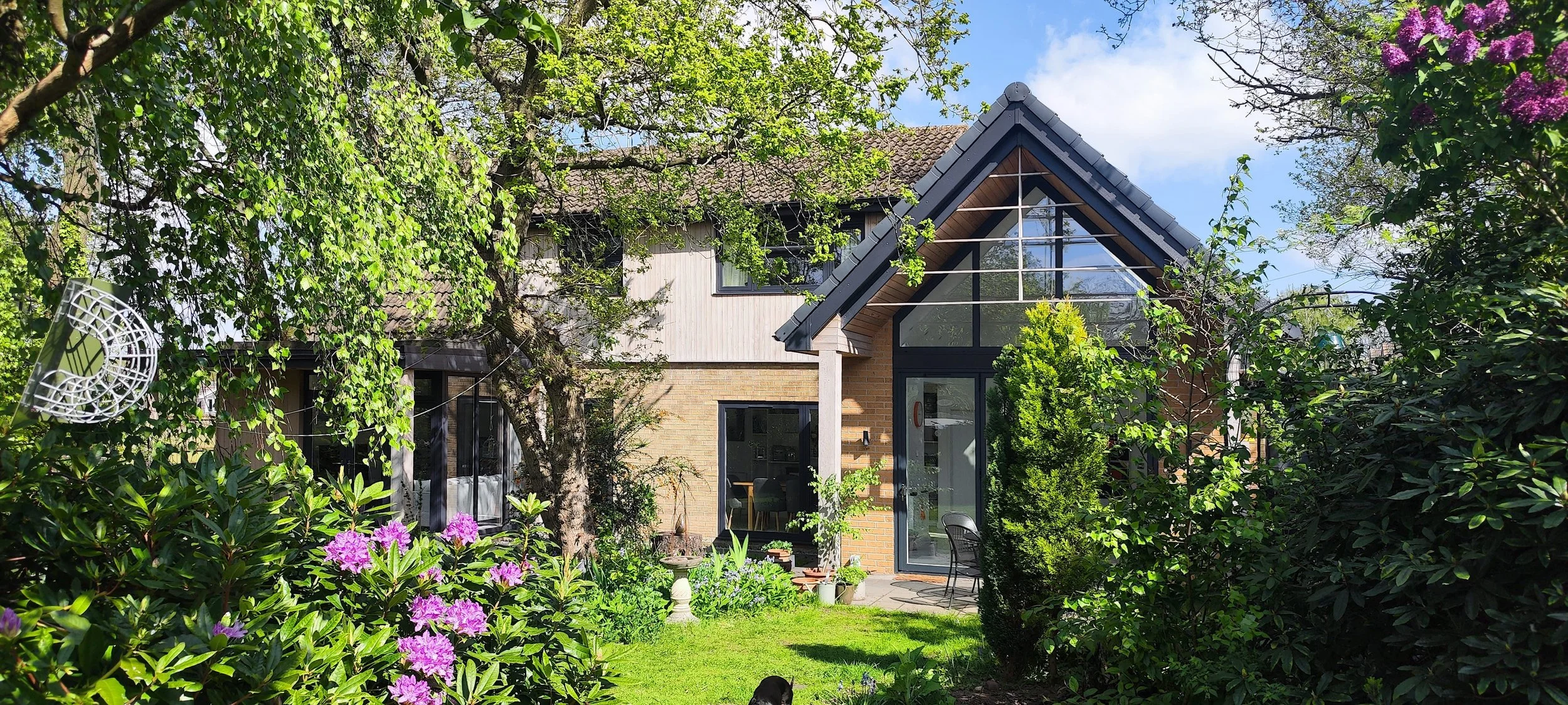 A modern two-story house with large glass windows and a sloped roof, surrounded by lush green trees and flowering plants in a garden.