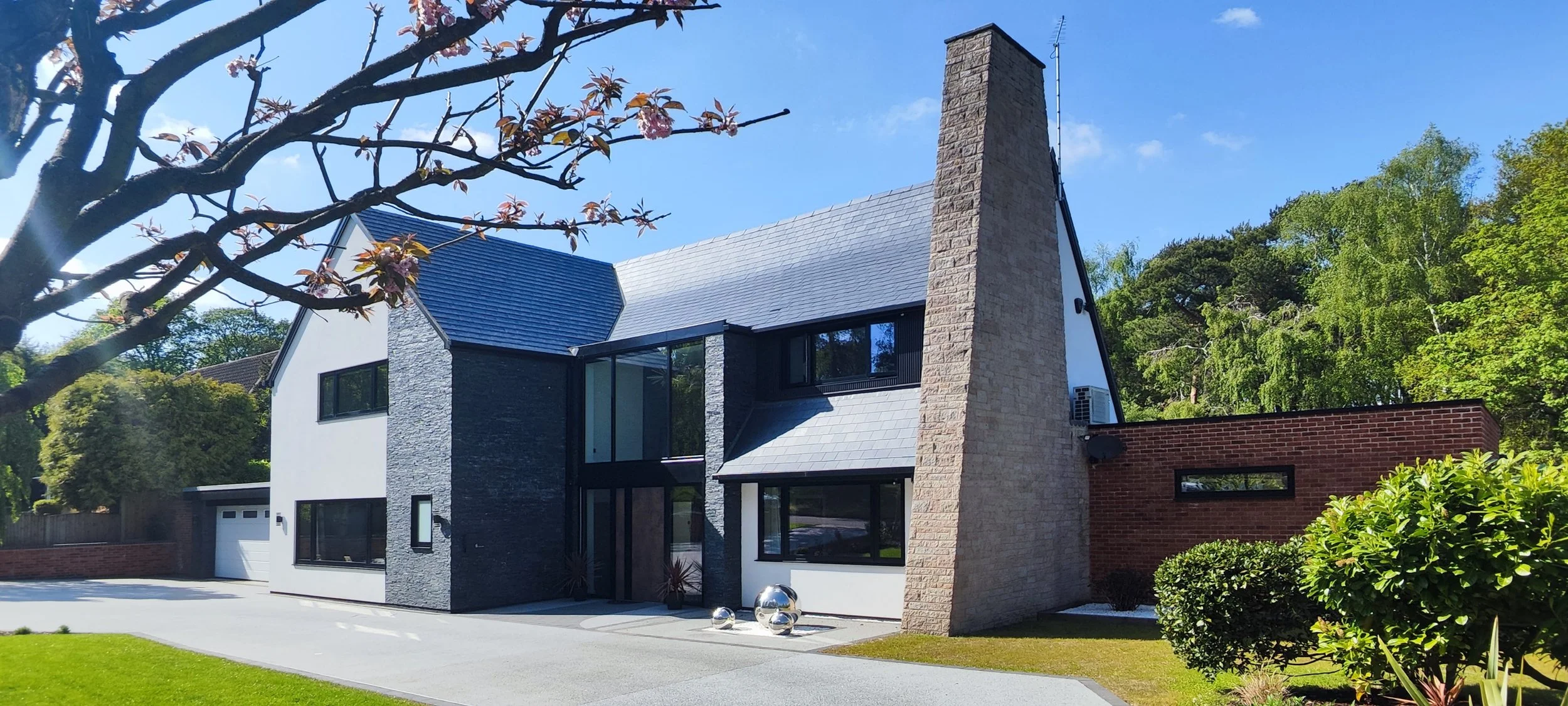 Modern two-story house with a mix of brick, dark gray stone, and white walls, large windows, a sloped roof, and a small front yard with trees and shrubs, under a blue sky.