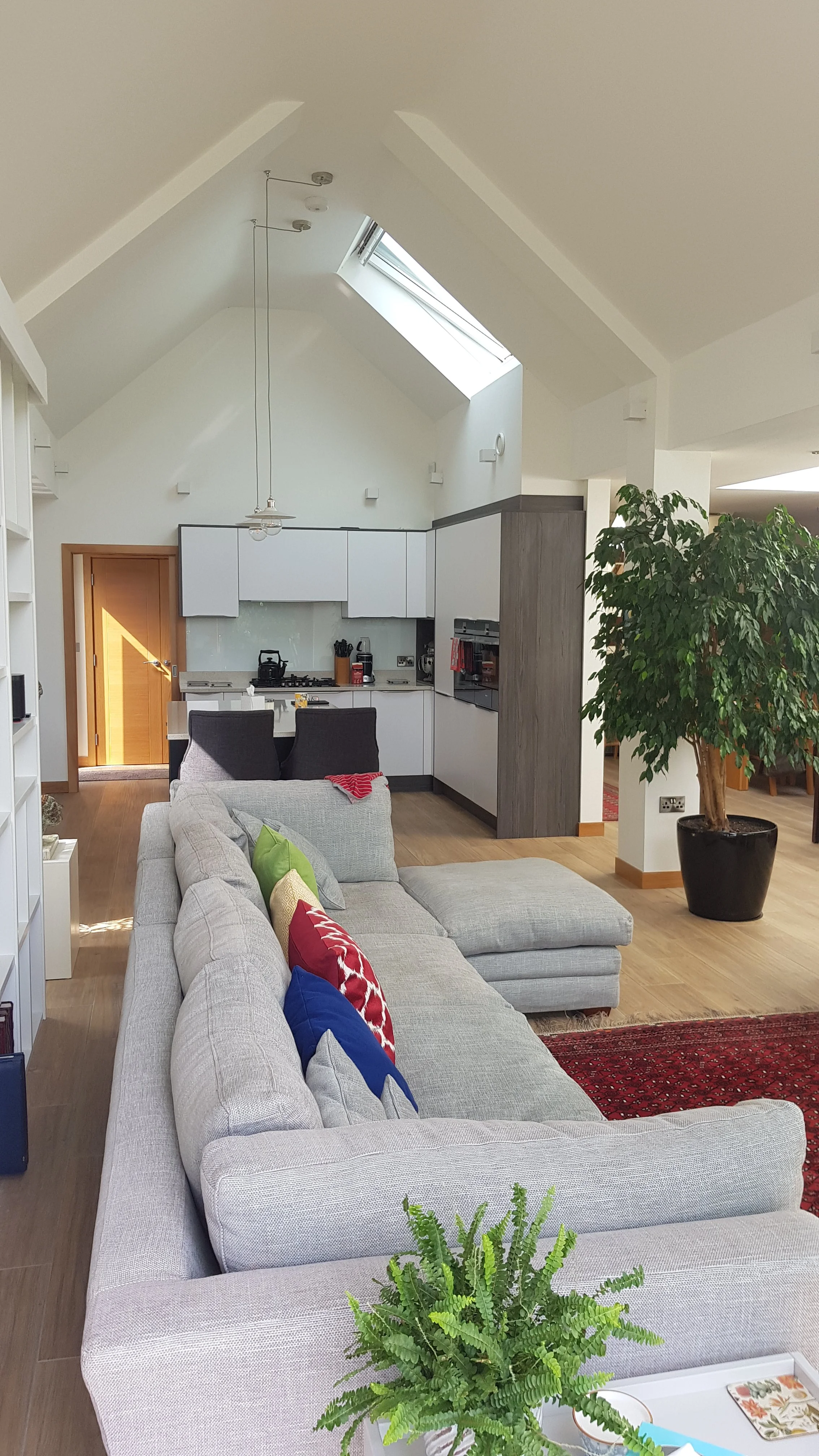 Interior of a modern living room with a light-colored sectional sofa with colorful pillows, a large potted plant, a partial view of a kitchen with white cabinets and a skylight, and a staircase leading to an upper level.