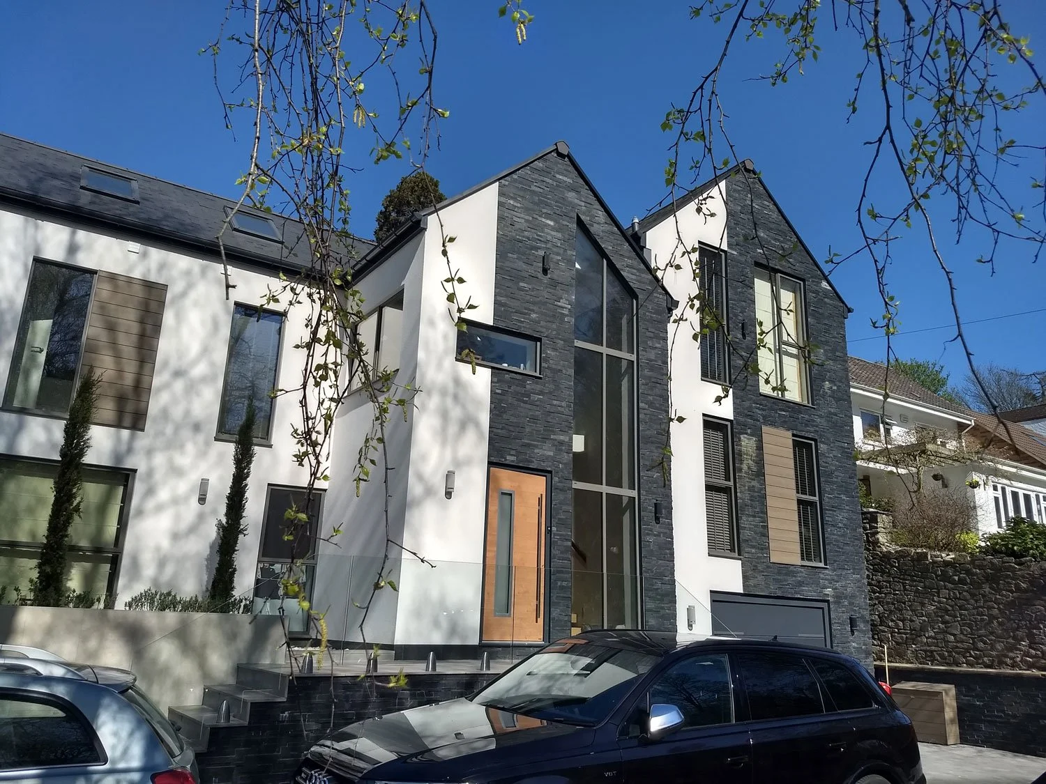 Modern house with black and white exterior, large windows, and wooden accents, parked cars in front, and a clear blue sky with tree branches overhead.