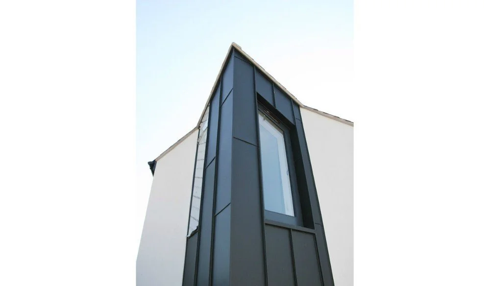 Close-up of a modern building's exterior corner with dark gray metal panels and a large window, against a clear blue sky.