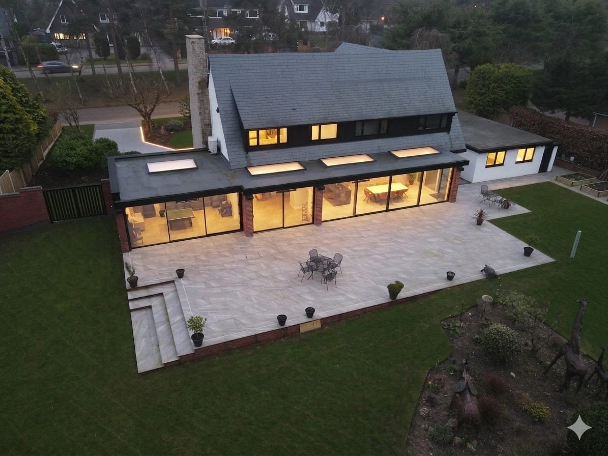 A modern house with large glass windows and a tiled outdoor patio surrounded by a green lawn and garden.