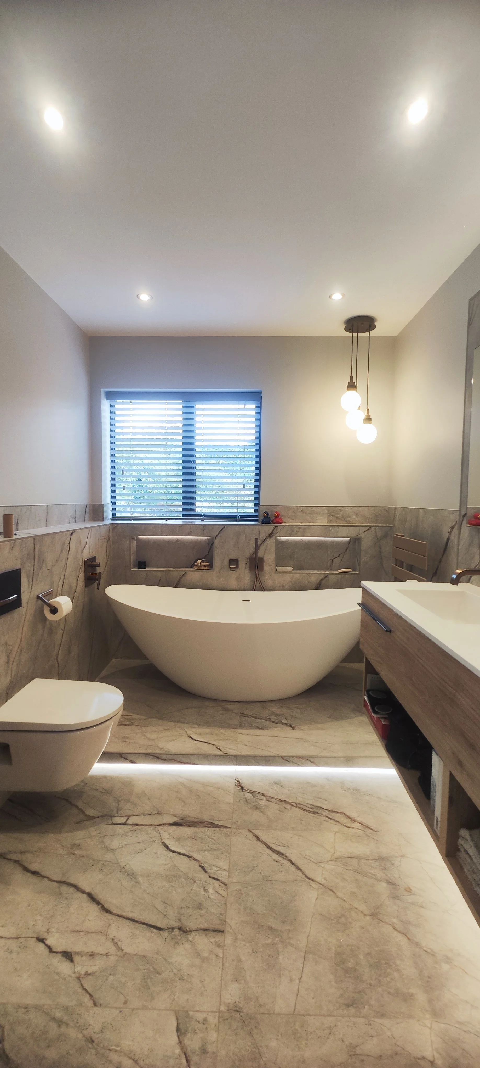 Modern bathroom with marble flooring and walls, a freestanding bathtub, a wall-mounted toilet, a window with blinds, and a vanity with a sink, illuminated by ceiling and pendant lights.