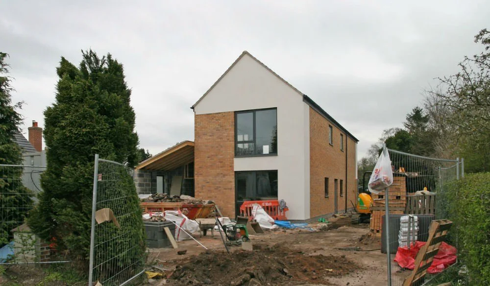 Under construction modern two-story house with brick and white siding exterior, construction materials and tools scattered in front, temporary fencing surrounding the site, trees and cloudy sky in the background.