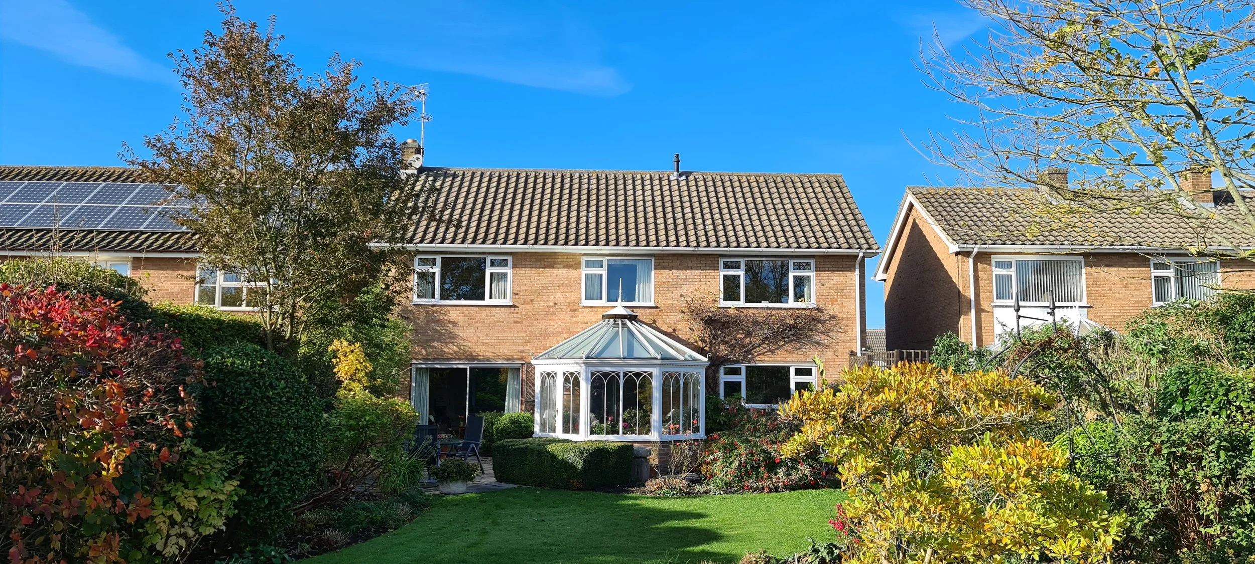 View of a suburban house with large windows, a glass conservatory, and solar panels on the roof, surrounded by a lush garden with trees and colorful bushes, under a bright blue sky.
