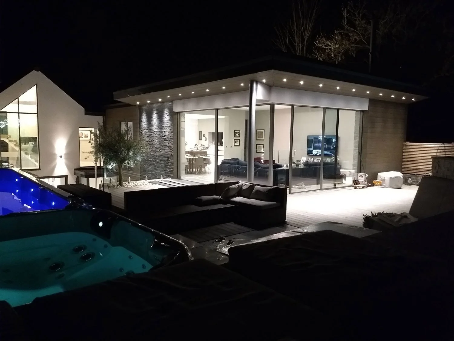 Night view of a modern house with large glass windows, outdoor seating, a hot tub, and a swimming pool with blue lighting.