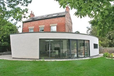 Modern white curved house with large glass windows in a grassy yard, with a red brick house and trees in the background.