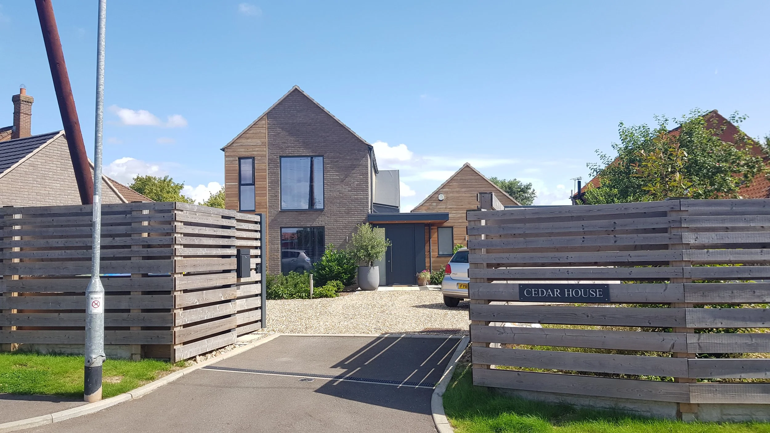 A modern house with a wooden facade, large windows, and a flat roof, surrounded by a gravel driveway and landscaped garden with potted plants and small trees. The house is fenced off with a wooden slat fence, and a small sign reads 'CEDAR HOUSE'. The