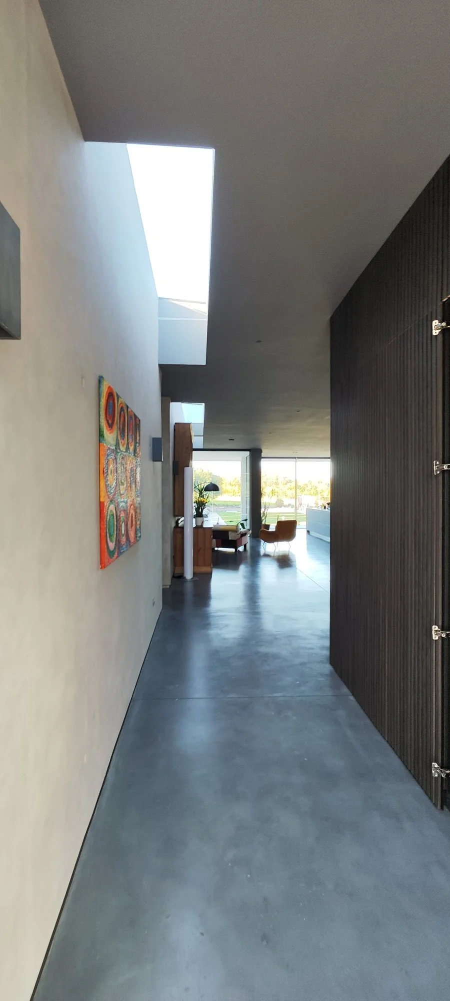 Modern hallway with polished concrete floor, beige wall with colorful artwork, and black wooden sliding door leading into a living space with large windows, a chair, and a view of trees outside.