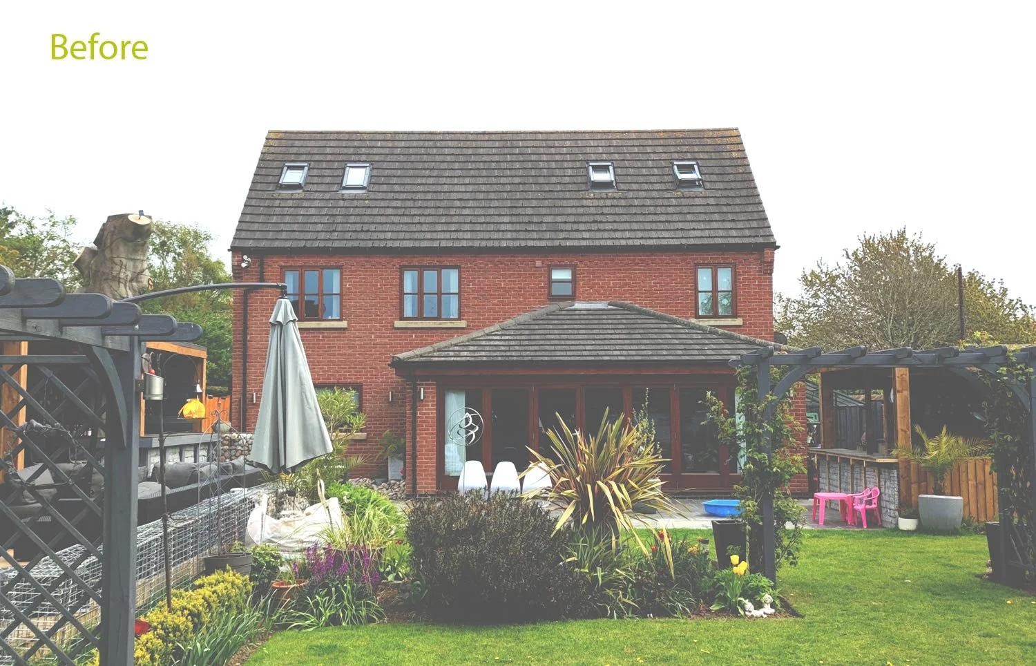 Rear view of a red brick house with a garden in the foreground, including a patio area, plants, and outdoor furniture.