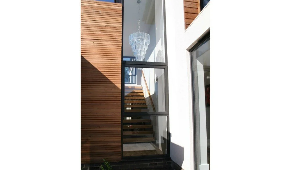 Exterior view of a modern house with wooden and glass elements, showing a staircase through a large window and a chandelier inside.