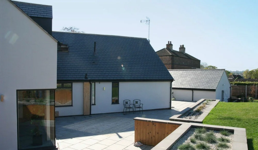 Modern backyard with paved patio area, two chairs, and raised planting beds, attached to a white house with a dark gray roof.