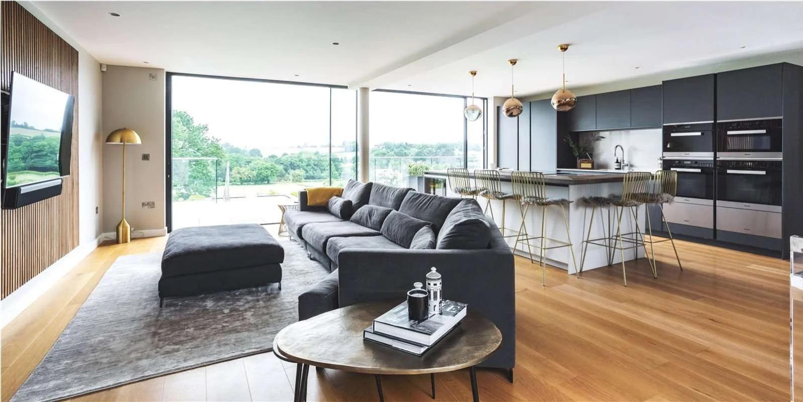 Modern open-concept living room and kitchen with large windows, gray sofa, wooden flooring, and sleek black kitchen cabinets with a white island and gold bar stools.
