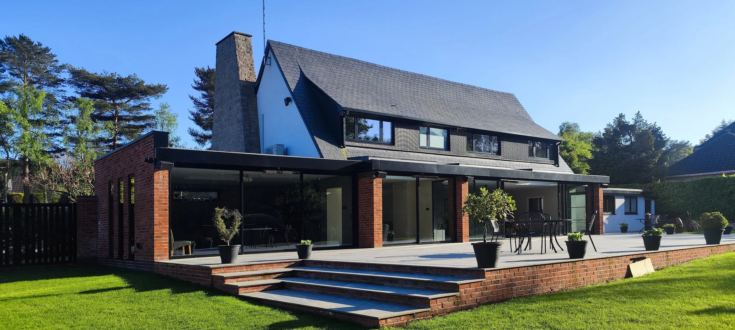 Modern house with a brick and glass outdoor patio, potted plants, and surrounding trees, under a clear blue sky.