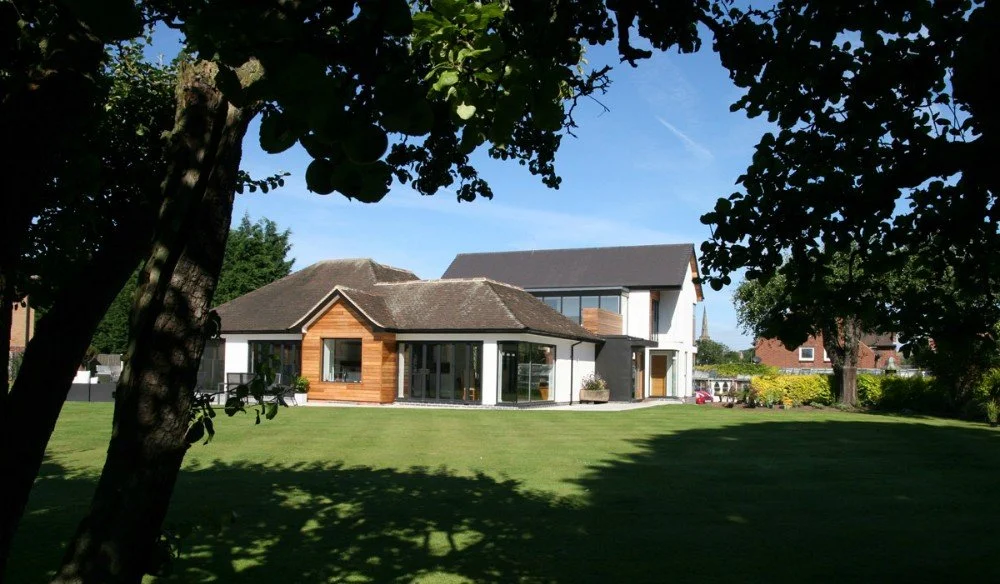 A modern two-story house with large windows and a mix of wood and white exterior, surrounded by a lush green lawn and trees, under a blue sky with a few clouds.